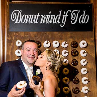 Bride kissing the grooms cheek while standing in front of a donut wall.