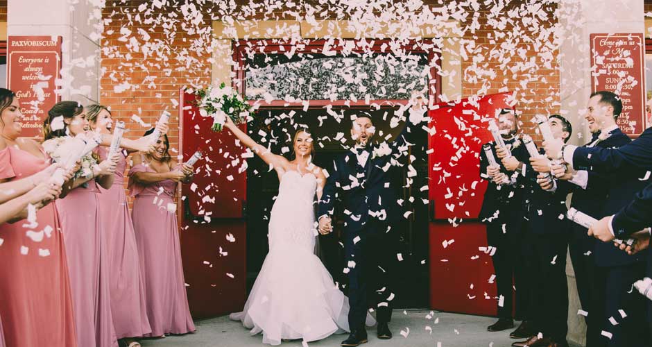 Bride and Groom have hands up in the air as they exit their church with confetti surrounding them.
