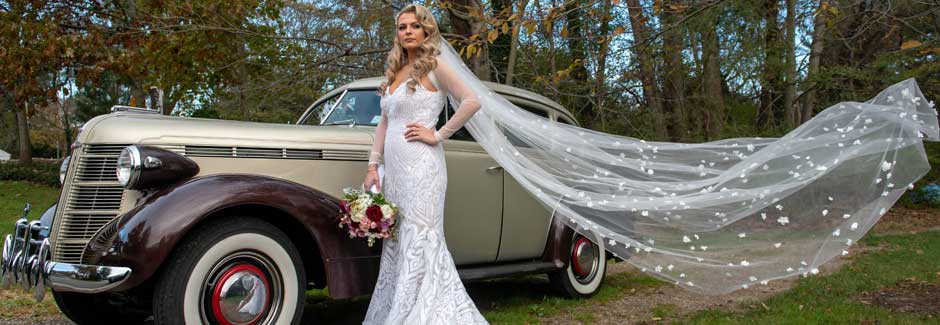 Bride holding her bouquet standing next to a Vintage Rolls Royce with her veil flying in the wind. 