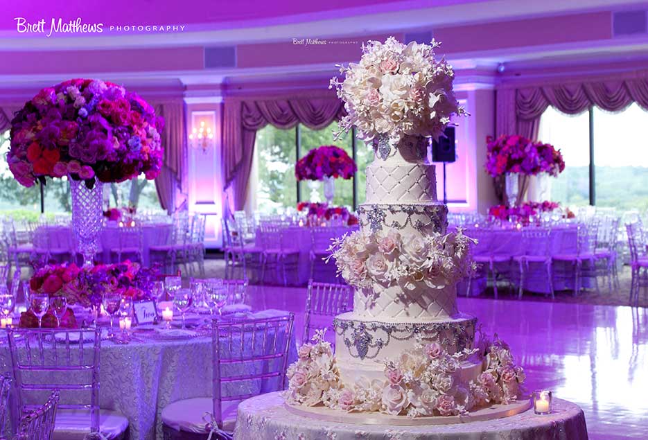 Close up of wedding cake surrounded by tables and purple up lighting.
