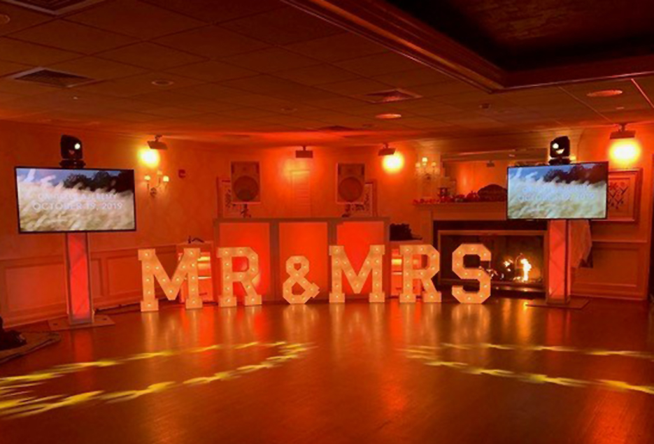 DJ setup in ballroom with orange uplighting and LED Mr. and Mrs. signs.