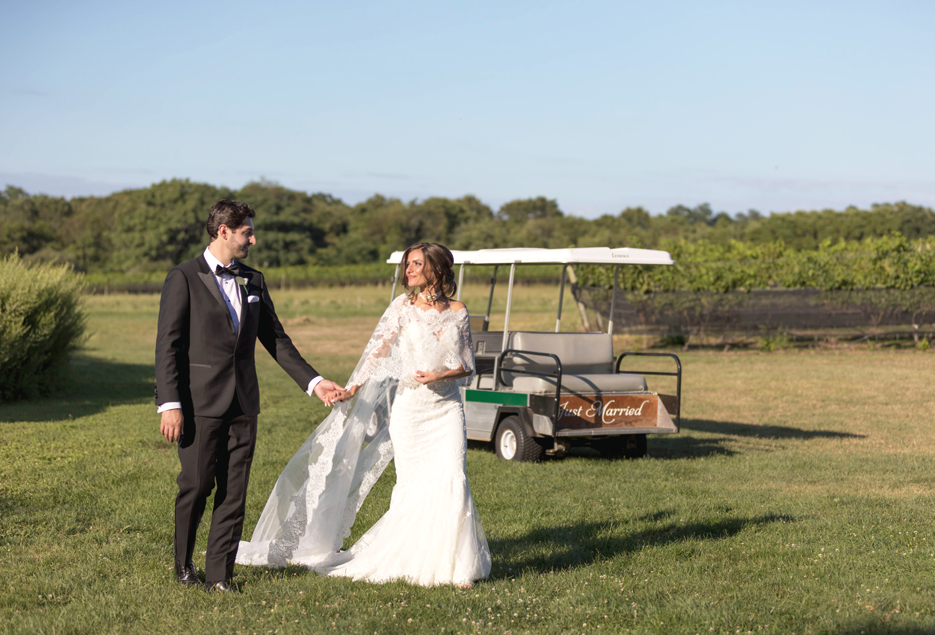Bride and groom hold hands and walk in a field front of a golf cart.