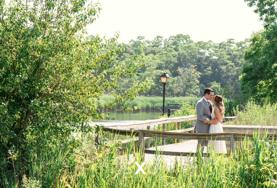 Bride and Groom kissing  outside while standing on wooden bridge overlooking lake. 