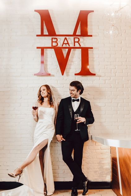 Bride and groom standing under a bar sign holding wine glasses. 