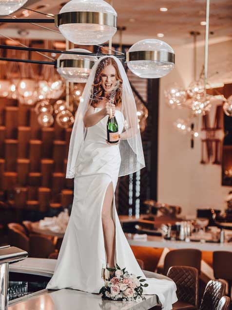 A bride holding a champagne bottle while standing on a bar. 