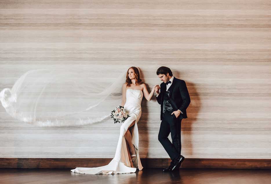 Groom holding the brides hand while posing in front of a wall. 