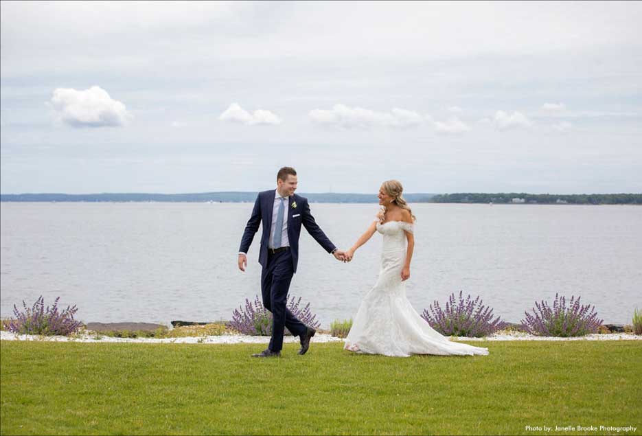 Groom and his bride walking on the grass along the water .