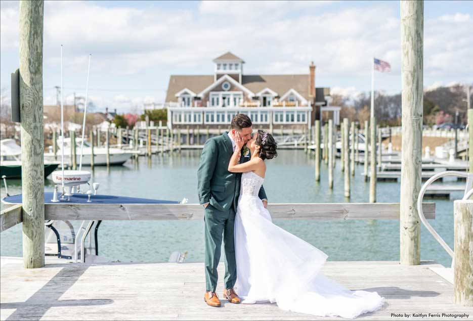Bride and groom kissing by the water with Peconic Bay Yacht Club in the background. 