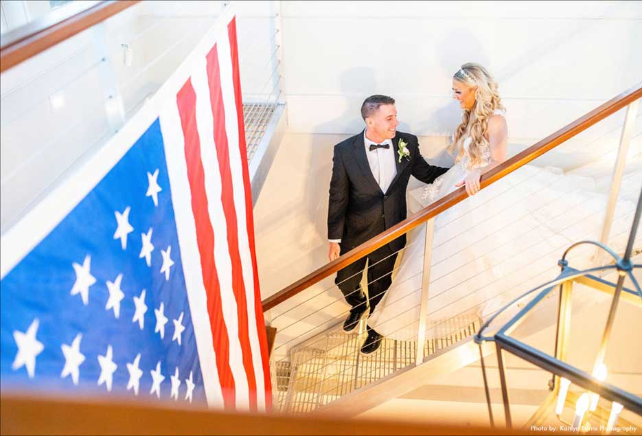 Bride and groom on staircase with the American flag hanging from ceiling. 