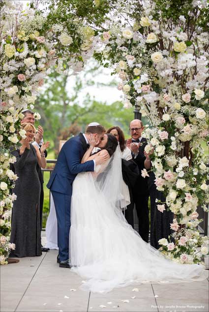 Bride and groom kissing under a flower Chuppah after exchanging their vows. 