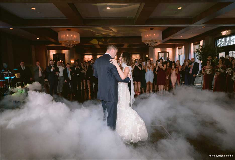 Couple dancing their first dance with white smoke mist surrounding them in front of guests. 