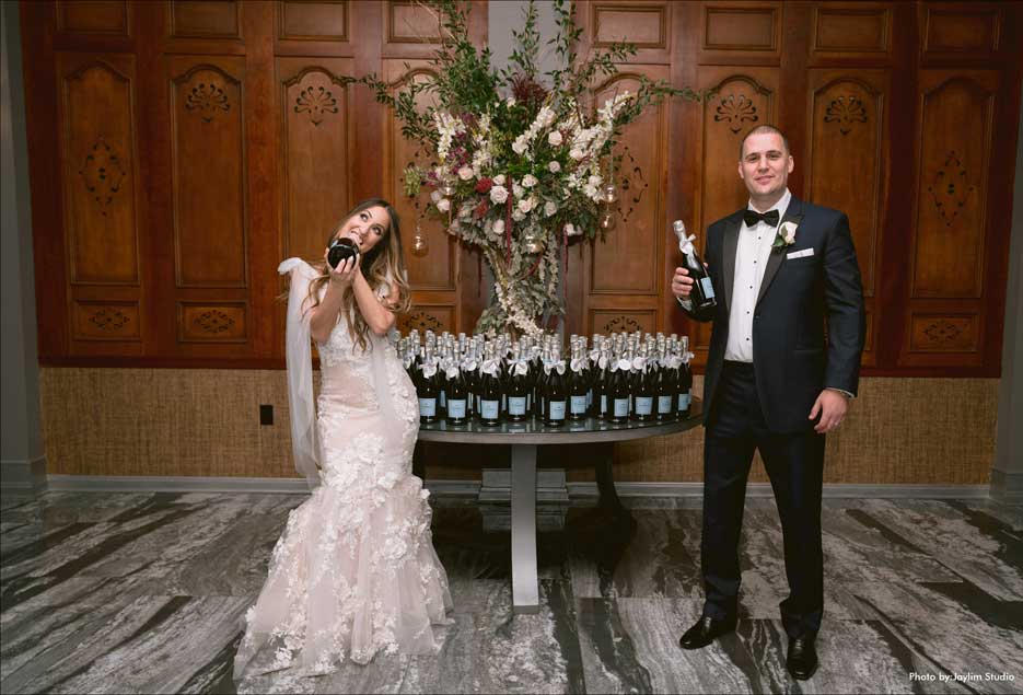 Bride and groom each holding a bottle of  champagne in front of their favor table. 